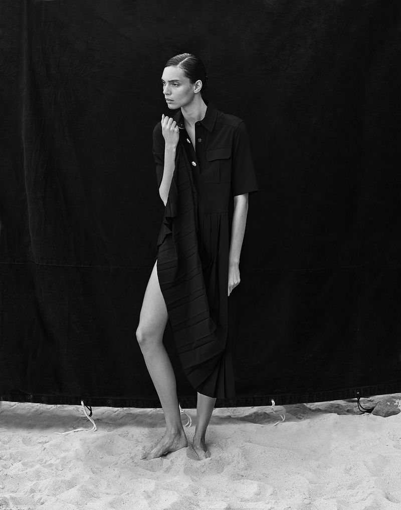 Monochrome studio shot of an adult female fashion model wearing a black short-sleeve dress, posing barefoot on a sand surface.