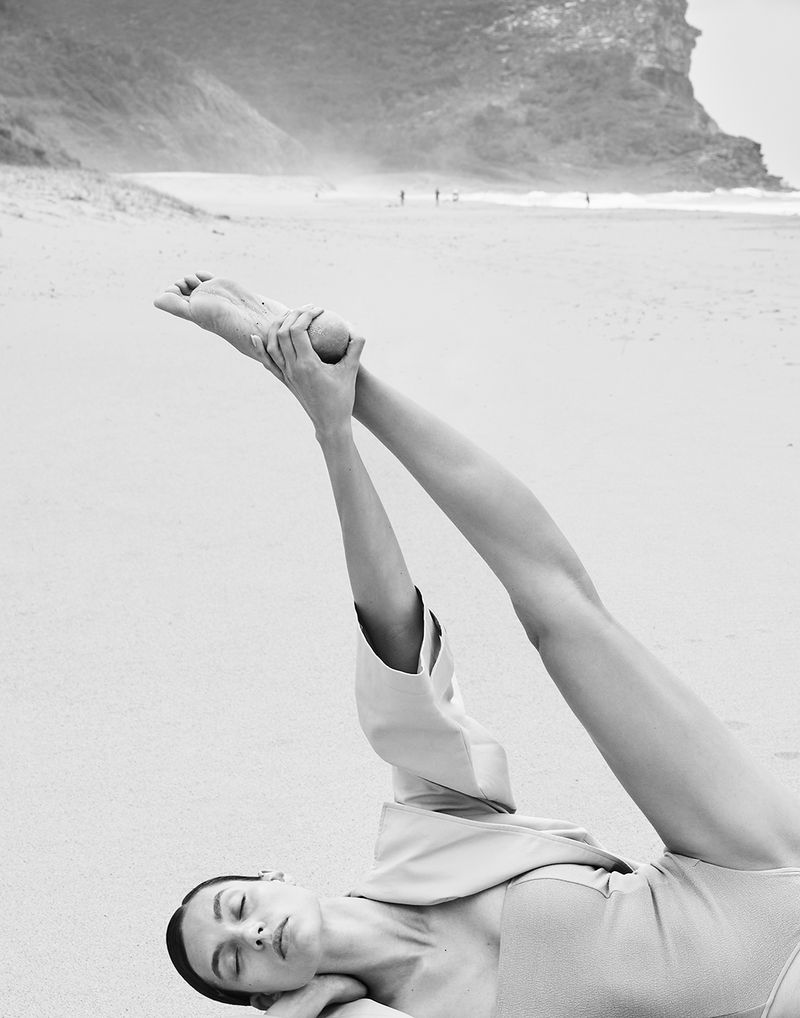 Black-and-white shot of a woman lying on a sandy beach with legs raised, holding a round object in her hands against an ocean backdrop.