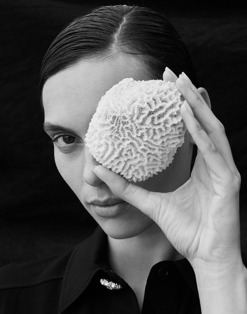 Black and white portrait of a woman holding textured coral over her eye, wearing a button-down shirt with a sparkling brooch.