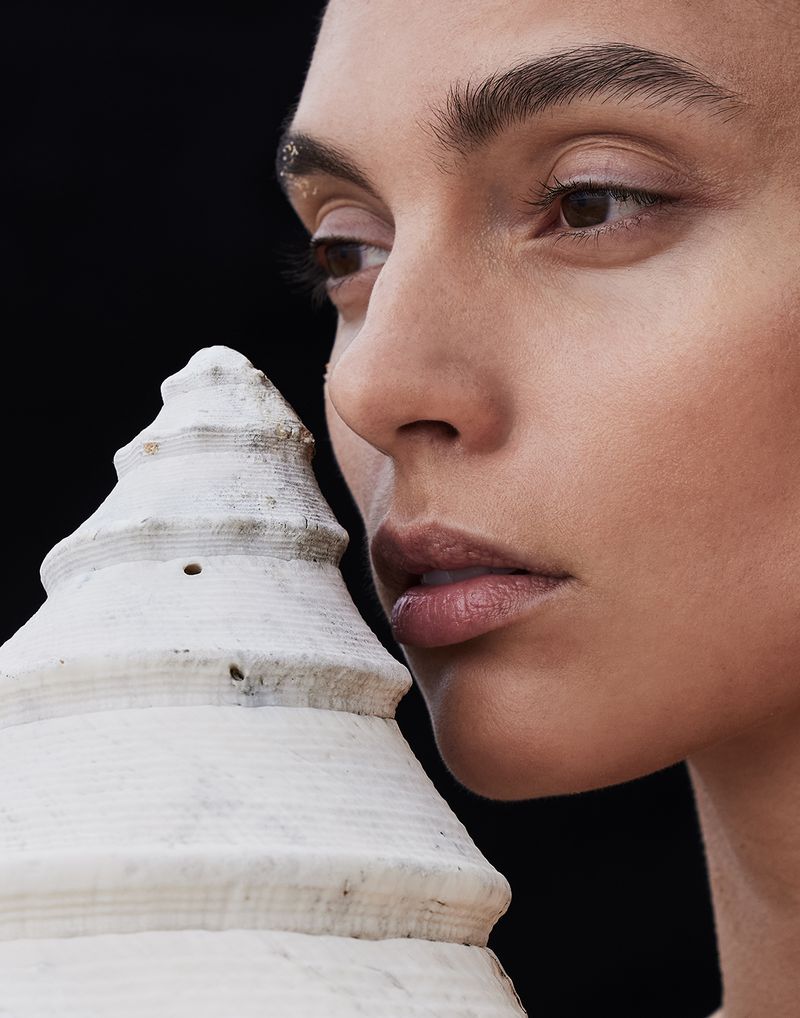 Close-up studio shot of a young adult female model with neutral expression posing with a decorative white seashell on a black backdrop.