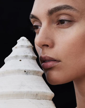 Close-up studio shot of a young adult female model with neutral expression posing with a decorative white seashell on a black backdrop.