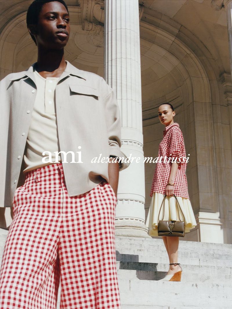 Two fashion models pose in front of classical architecture. One model wears a beige jacket and red checkered pants, the other a checkered dress.
