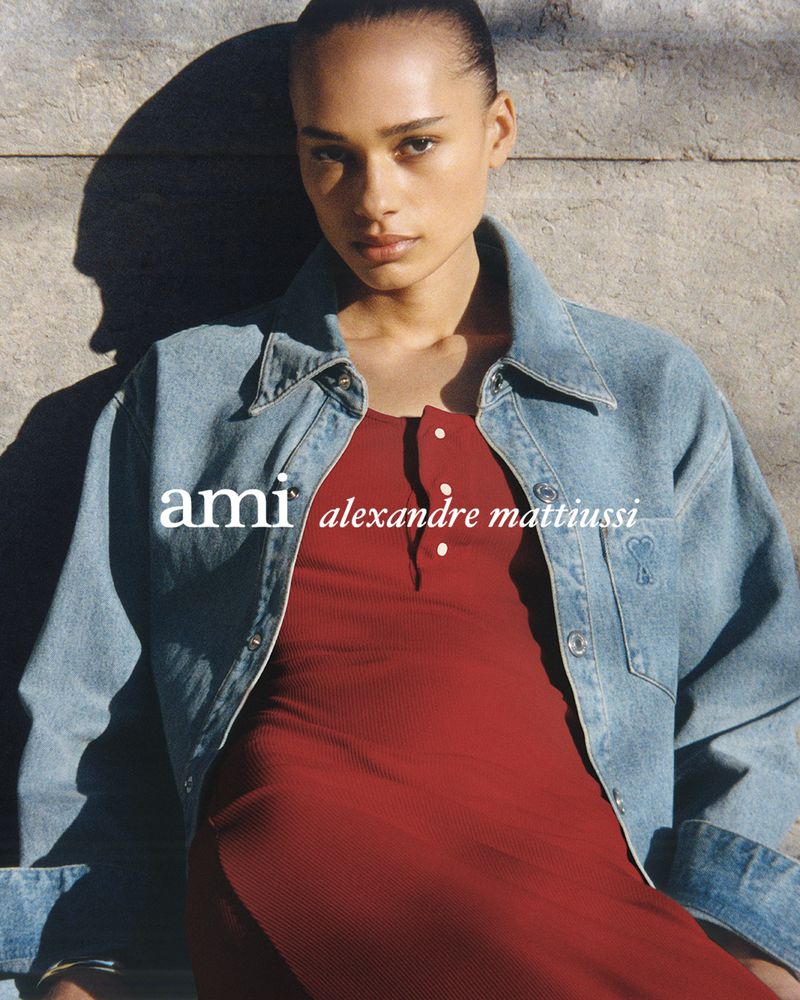Portrait of a woman in a red dress and denim jacket, posing against a wall in diffused sunlight, with the 'ami alexandre mattiussi' logo.