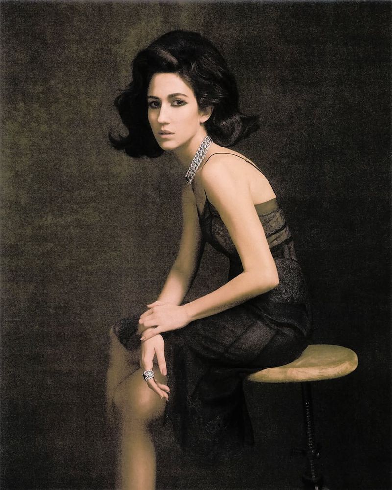 Portrait of a woman with dark hair and a black dress sitting on a stool in front of a textured background in a photo studio.
