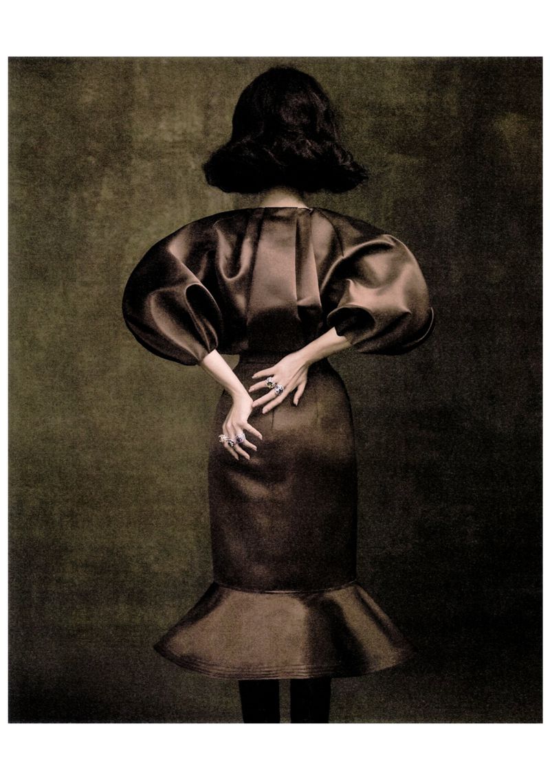 Back view of a stylish adult woman in a brown satin dress with puffy sleeves and a flounce hem. She is wearing rings and posing indoors.