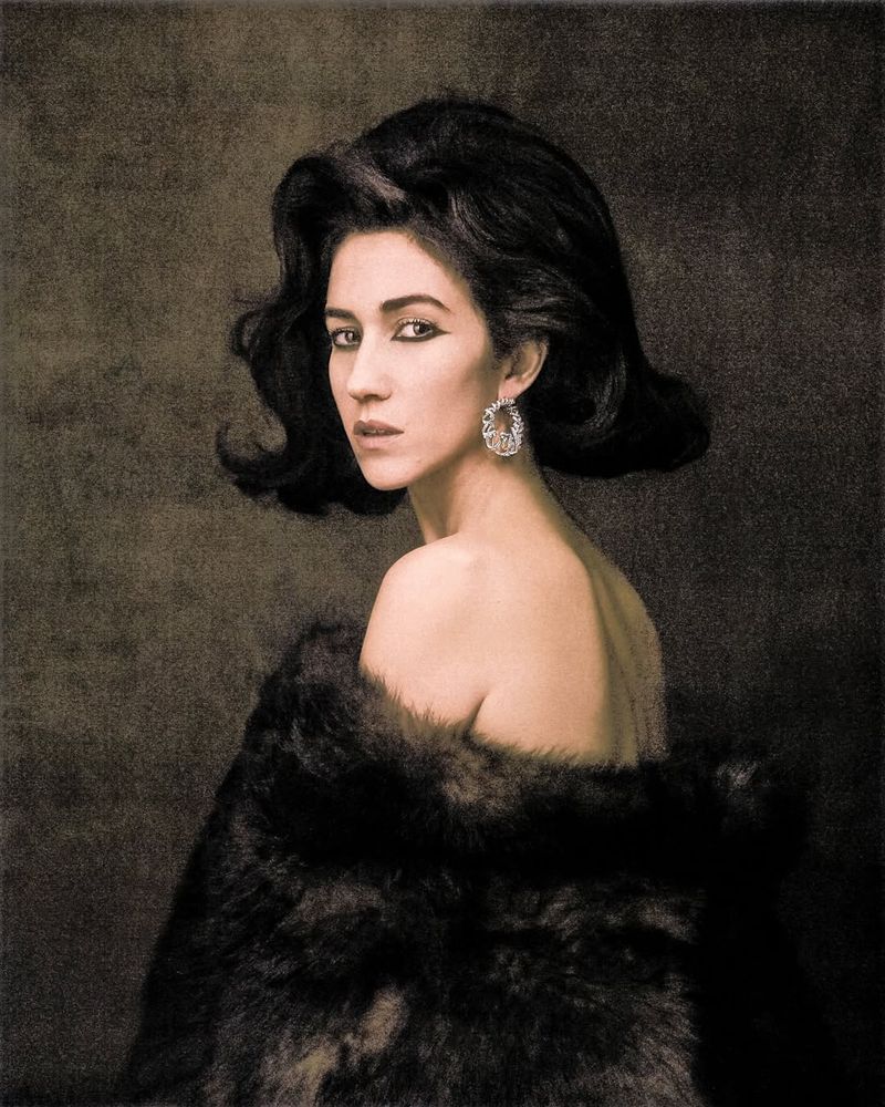 Portrait of an adult female with dark hair, wearing a fur coat and earrings, looking over her shoulder in front of a neutral background.