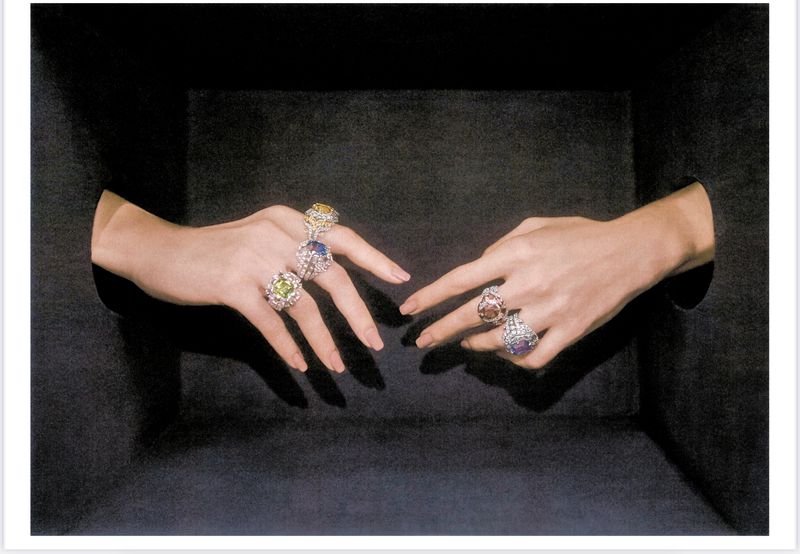 Close-up of two adult female hands adorned with rings featuring various gemstones, against a dark black background, jewelry advertisement.