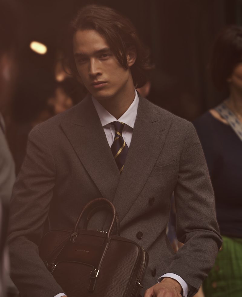 A fashionable adult male in a grey suit and tie holds a leather briefcase indoors with other adults blurred in the background.