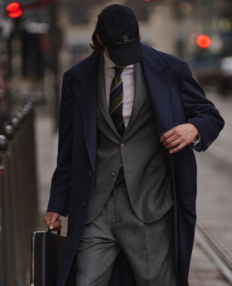 A man in a blue overcoat and baseball cap walks on a city sidewalk carrying a black briefcase, creating a business professional style.