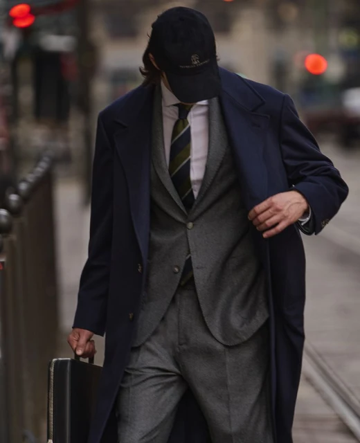A man in a blue overcoat and baseball cap walks on a city sidewalk carrying a black briefcase, creating a business professional style.