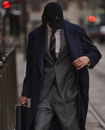 A man in a blue overcoat and baseball cap walks on a city sidewalk carrying a black briefcase, creating a business professional style.