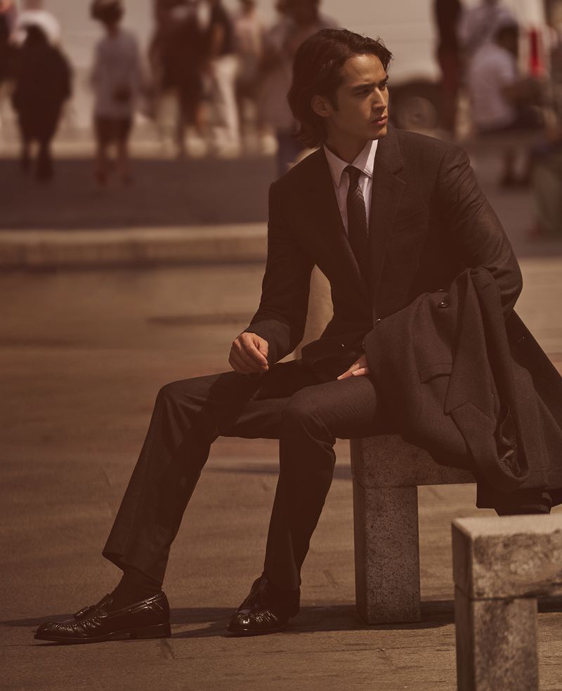 Man in a suit sits on a concrete barrier outdoors with blurred pedestrians in the background, fashion shot, warm color grading.