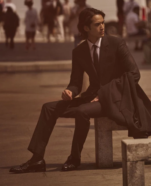 Man in a suit sits on a concrete barrier outdoors with blurred pedestrians in the background, fashion shot, warm color grading.