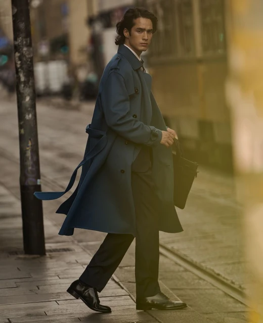 A brunette man in a teal trench coat and black dress pants walks across the street in a city while holding a black bag.