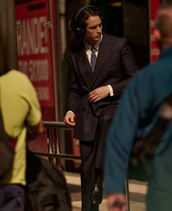 Man in a dark double-breasted suit wearing over-ear headphones, posing by an urban street railing between blurred pedestrians.