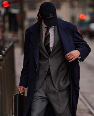 Man in a navy overcoat and grey suit walking down a city street while carrying a black briefcase.