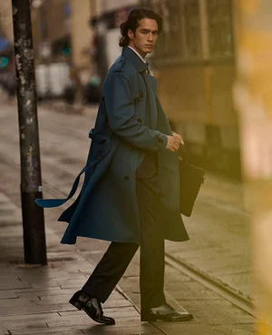 A man in a petrol blue trench coat walks along a city street carrying a black leather bag near tram tracks.