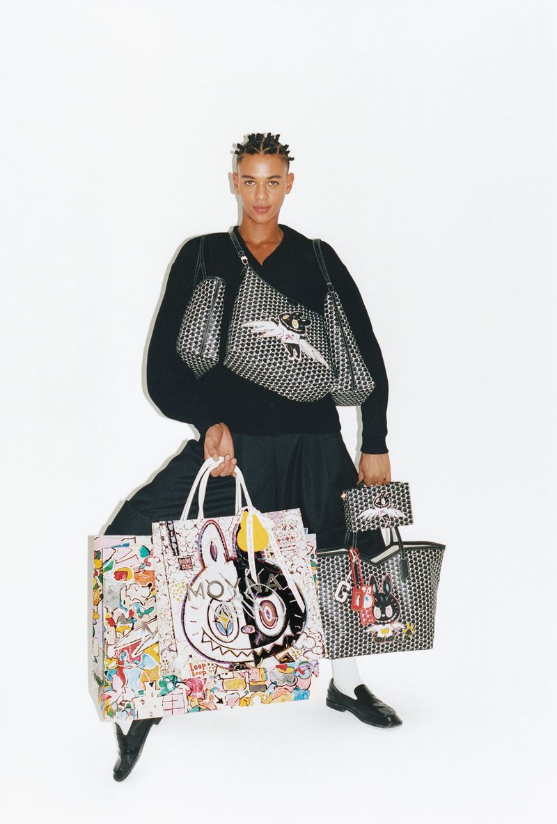 Guillaume Diop in black knitwear holding several printed Moynat x Kasing Lung tote bags against a seamless white background.
