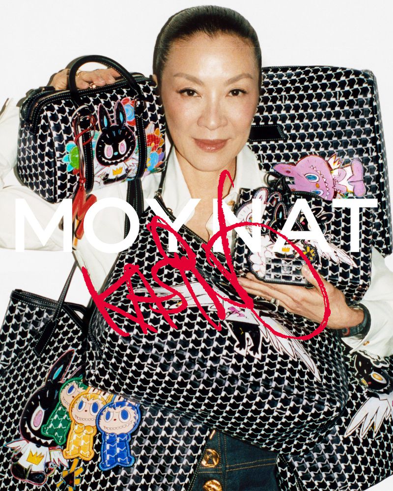 Michelle Yeoh poses with patterned handbags and colorful bag charms, indoors with a white background in a promotional studio shot.