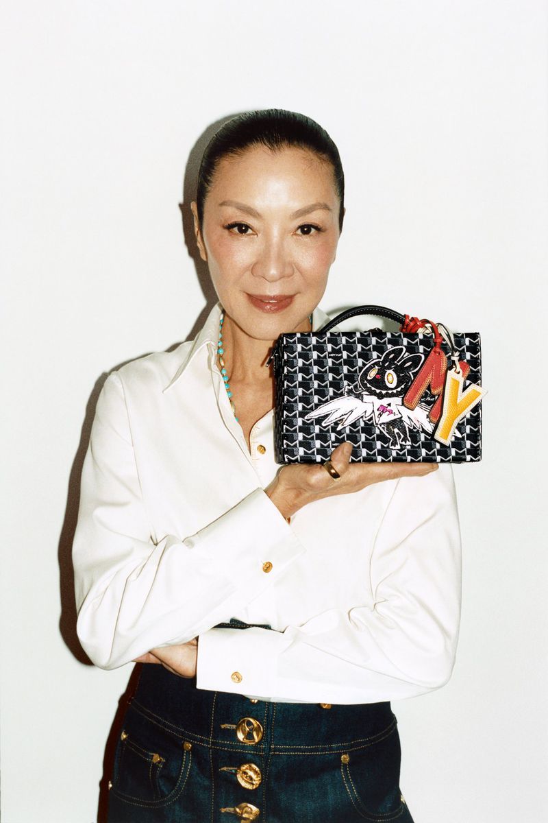 Michelle Yeoh presents a patterned handbag in a white shirt and dark skirt against a plain background, showcasing fashion.