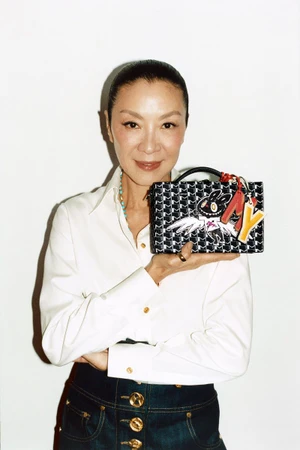 Michelle Yeoh holding a patterned Moynat box bag with charms against a plain white studio background.
