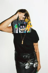 Carine Roitfeld holding an illustrated Moynat handbag over her face wearing a black t-shirt and leather skirt against a white wall.