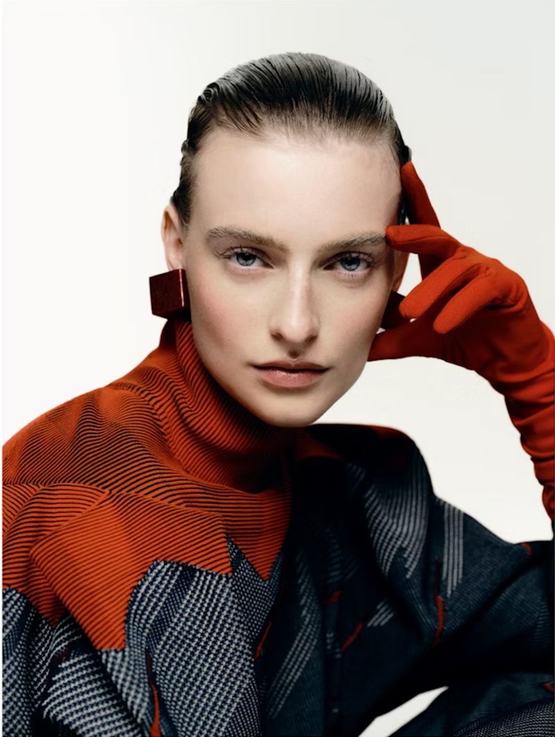 Fashion portrait of a woman in a geometric turtleneck and red gloves in a brightly lit studio with hair slicked back.