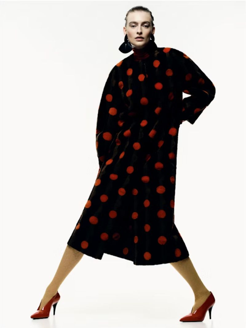 Elegant woman in a polka-dot coat and red high heels poses in a studio setting with a clean white backdrop, fashion editorial.