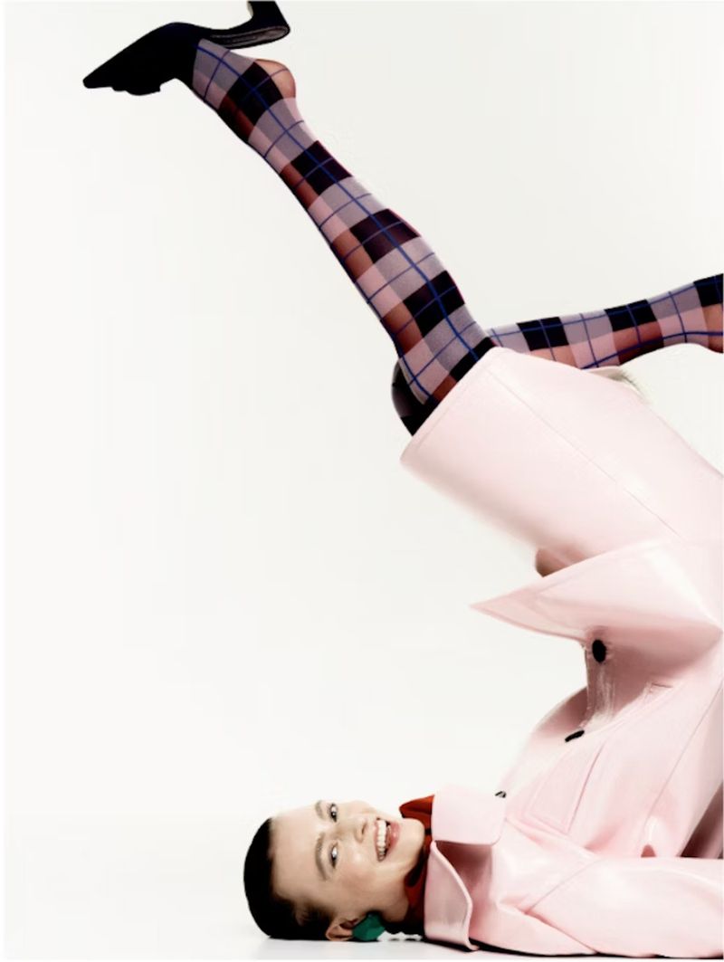 Woman in pink jacket and plaid tights poses creatively against a white background with her legs in the air and a smile on her face.
