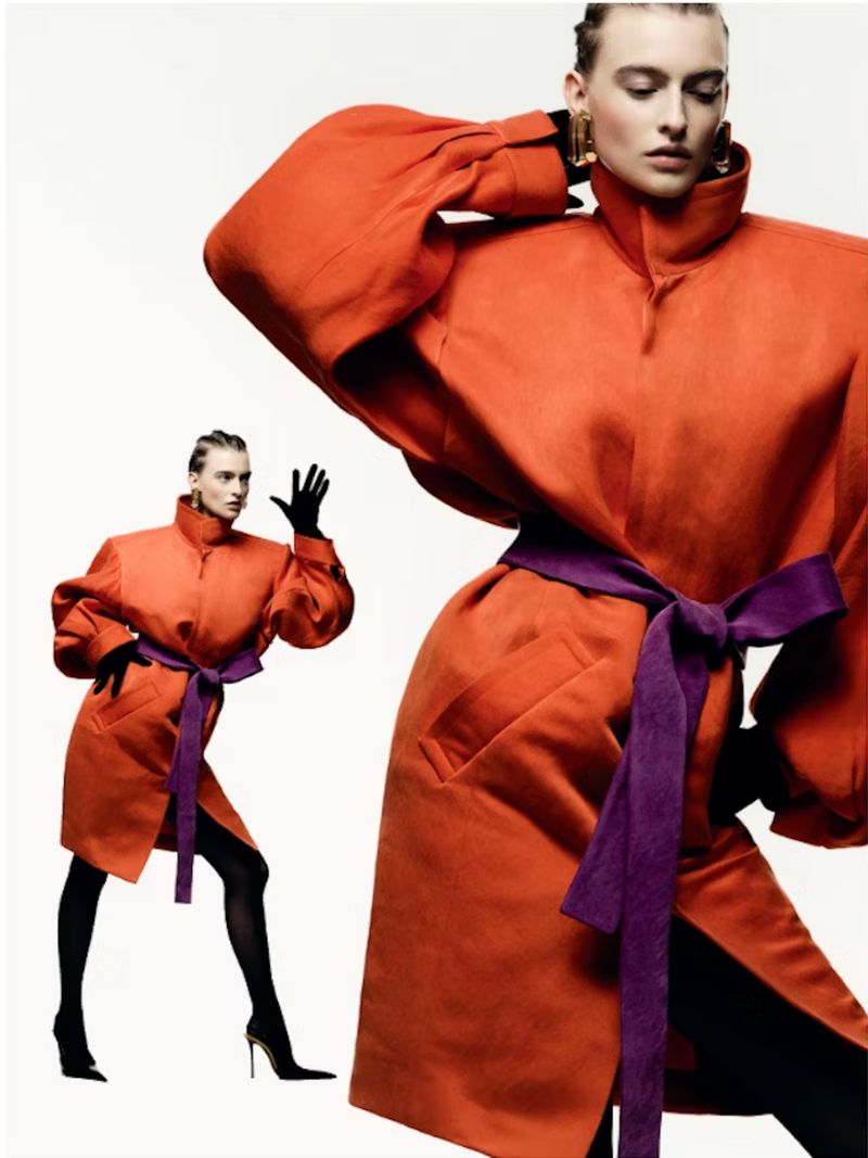 Fashion shot of a model wearing a large orange coat with a purple belt, black tights, and heels posing against a white backdrop.
