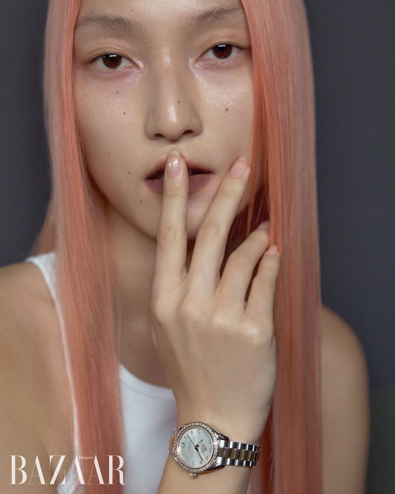 Close-up studio shot of an adult female fashion model with pale skin and salmon-colored hair, wearing a wristwatch, against a dark background.