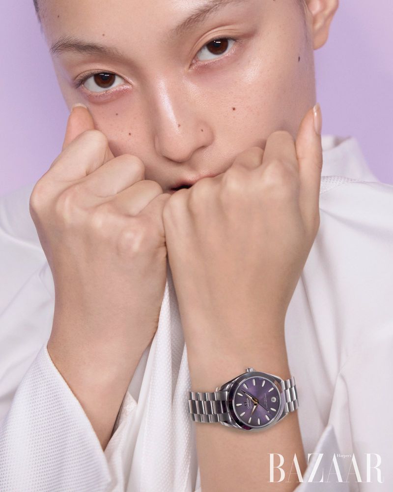 Close-up of a woman with fair skin in a white top, showing a silver wristwatch on her wrist against a light purple backdrop.