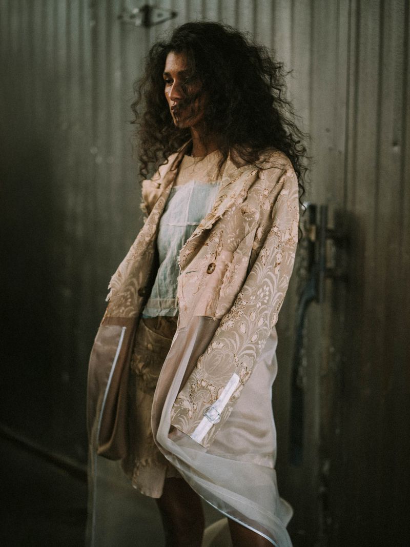 Woman in an embroidered tan blazer and shorts poses indoors in a fashion editorial-style shot against a textured wall.