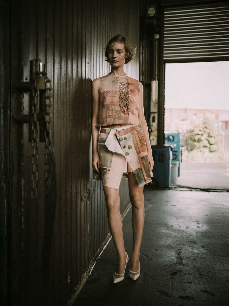 A fair-skinned woman posing in a fashion top and skirt, accessorized with high heels, stands in an industrial indoor location.