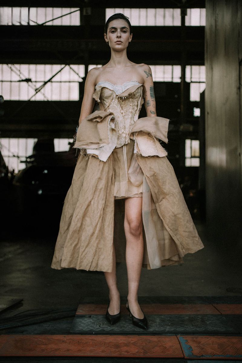 Full shot of a woman in a textured, strapless gown with beige tones, standing indoors with an industrial backdrop and dark shoes.