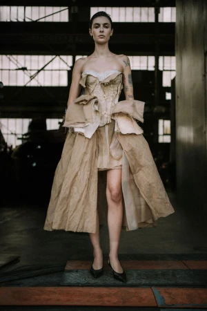 Full shot of a woman in a textured, strapless gown with beige tones, standing indoors with an industrial backdrop and dark shoes.