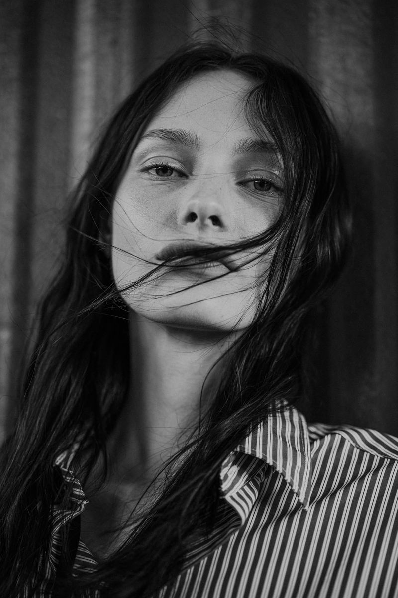 Monochrome portrait of a young woman with long dark hair, wearing a striped shirt, posing against a textured grey wall.