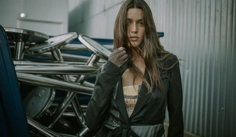 Adult woman with long brown hair in a jacket poses indoors near stacked silver metal tables and a grey metal wall in soft studio light.