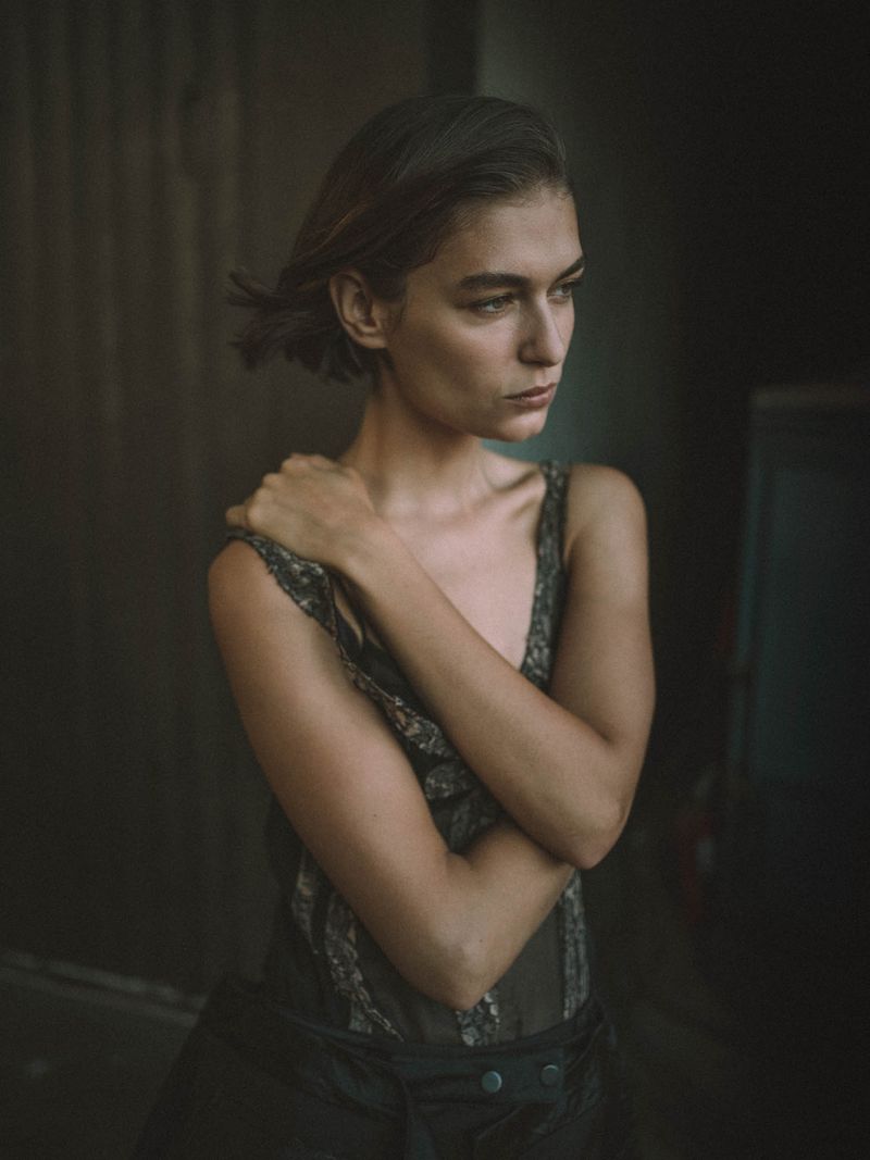 Portrait of a young woman with short brown hair and dark clothing, arms crossed, with a dark, shadowy background, neutral expression.