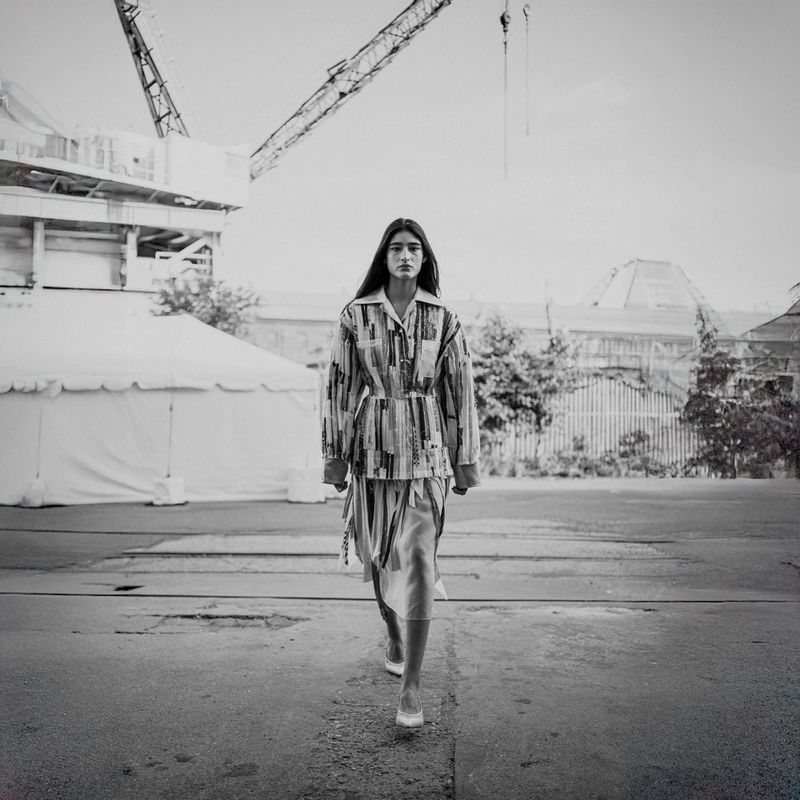 Runway fashion: Model presents striped shirt and skirt on industrial concrete, featuring crane, factory, and bright monochrome lighting.