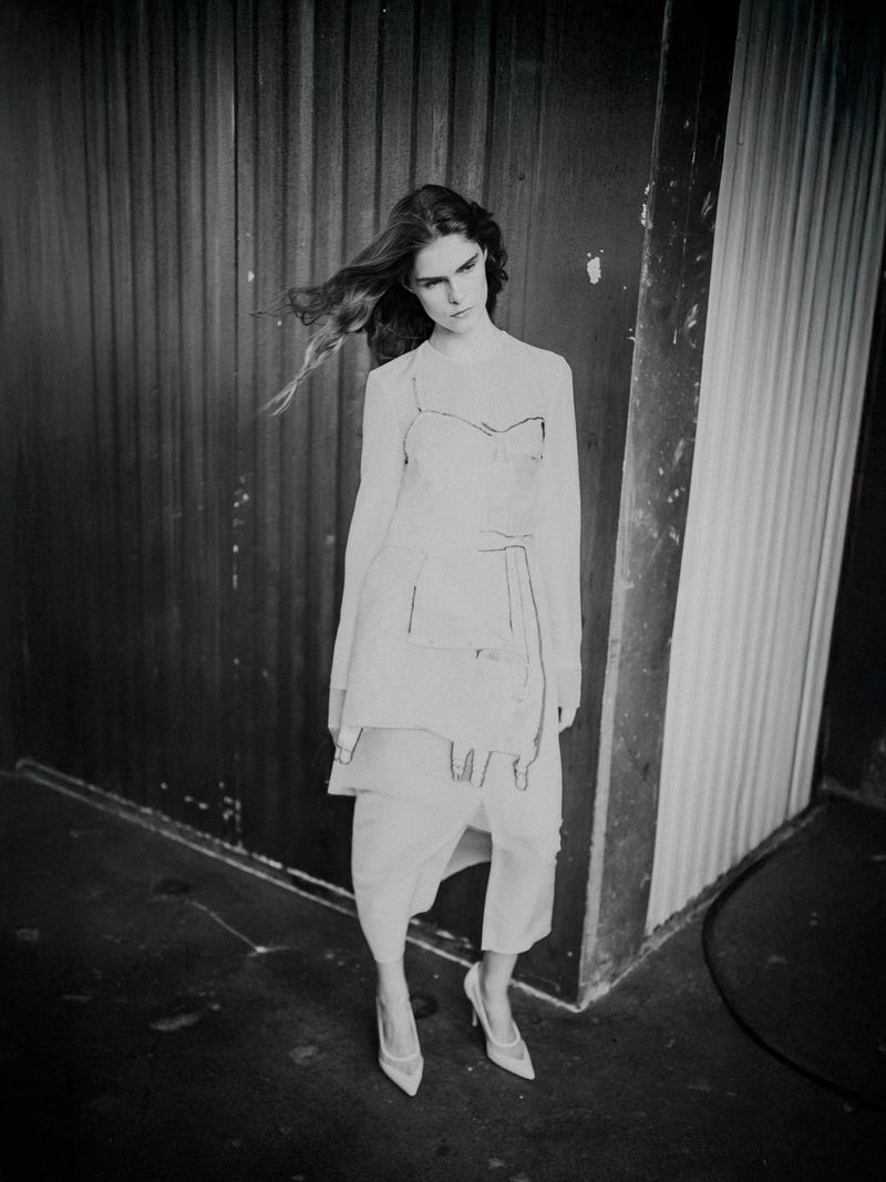 A woman in a white outfit and heels poses against a wall with peeling paint and a metal door in a high-contrast black and white shot.