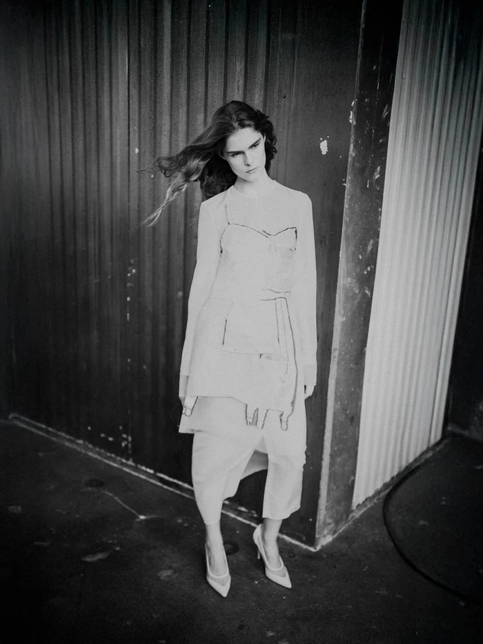 Model with long blowing hair wearing layered light-colored dress and heels standing against a textured industrial wall backdrop