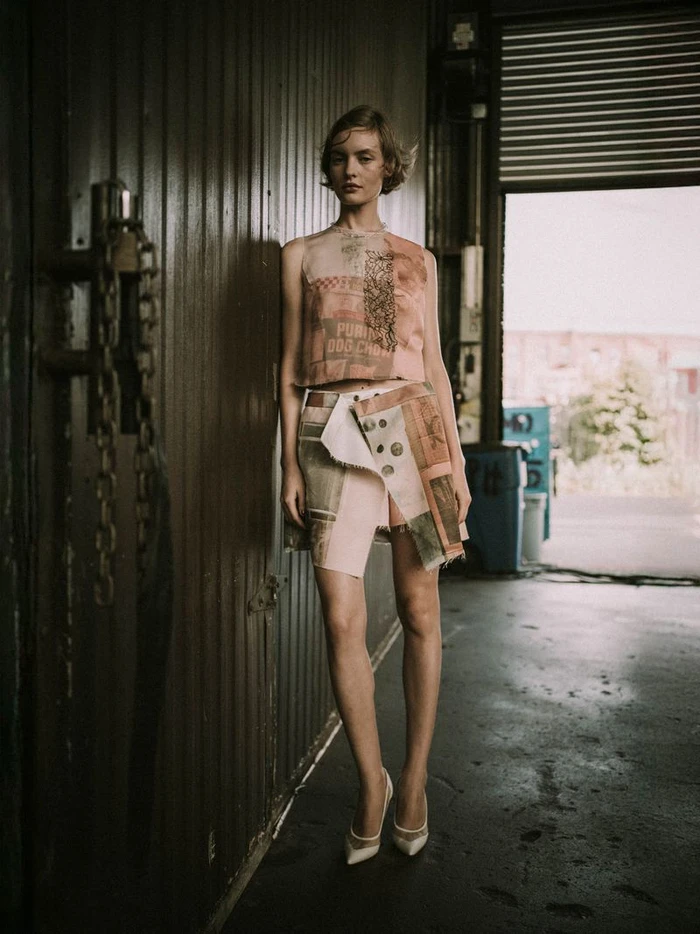 A fashion model in a graphic printed crop top and wrap mini skirt posing by a corrugated wall backstage at the Jason Wu SS26 show.