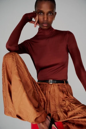 Fashion shot of a Black woman in a brown turtleneck top and wide, patterned brown pants, seated against a grey background.