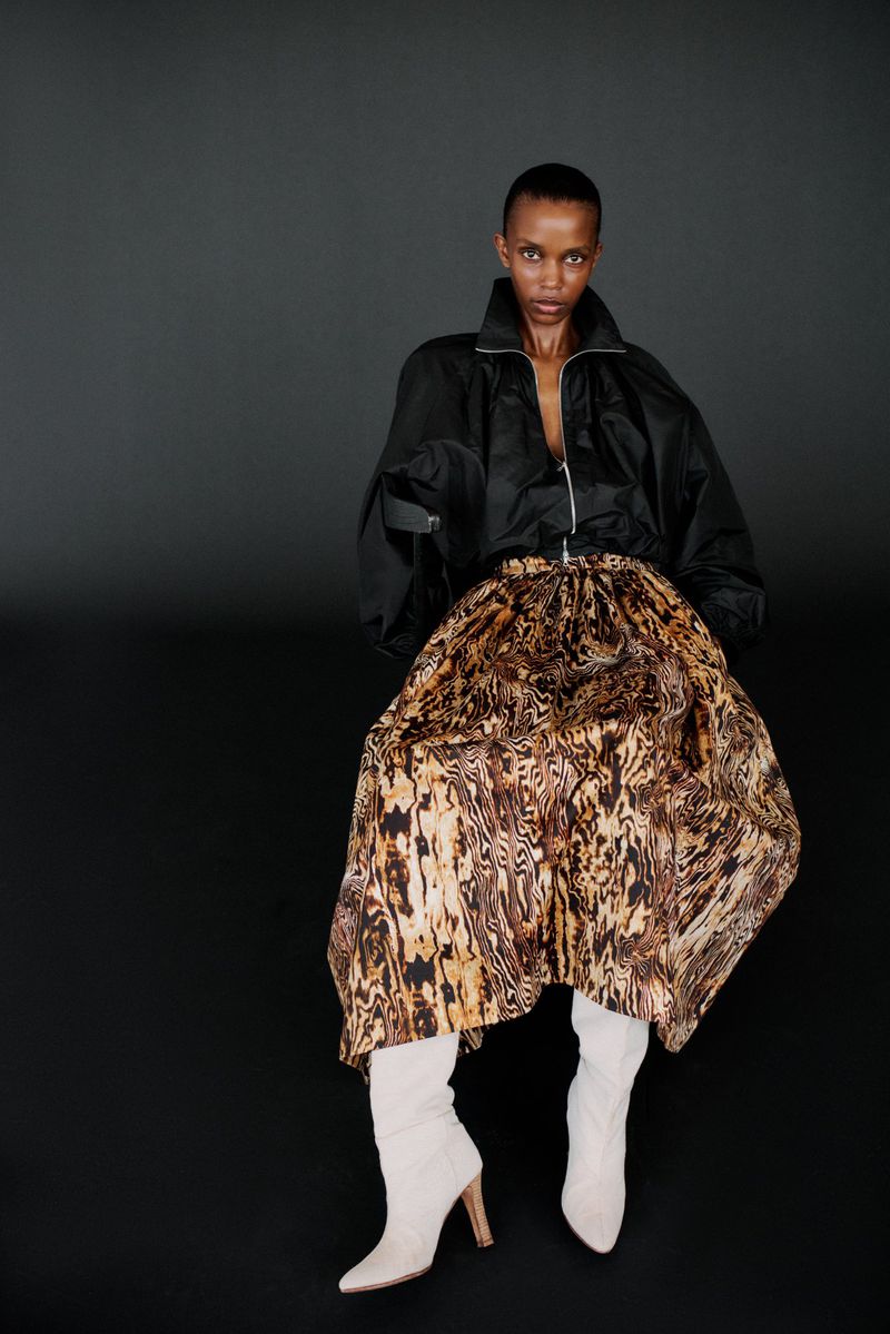 Woman in a black jacket and animal print skirt posing in a studio with a dark background. Neutral-colored, high-heeled boots complete the look.