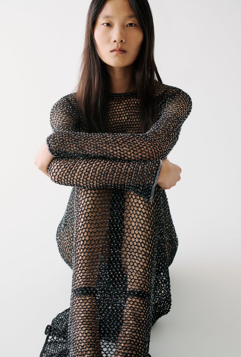 Asian woman in a black mesh dress, posing in front of a white background in a fashion studio, dark hair, arms crossed.