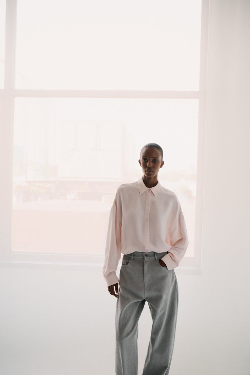 A woman with short hair poses in a pink button-down shirt and gray pants against a bright window, casting a soft, diffused light.