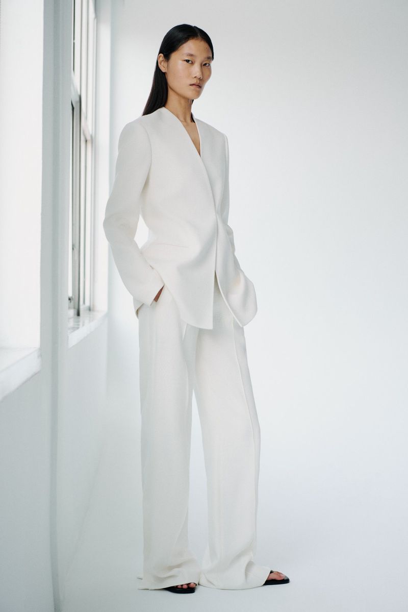A stylish Asian woman with long hair poses in a white suit with wide-leg trousers in a bright, minimally decorated studio setting.