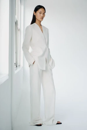 A stylish Asian woman with long hair poses in a white suit with wide-leg trousers in a bright, minimally decorated studio setting.