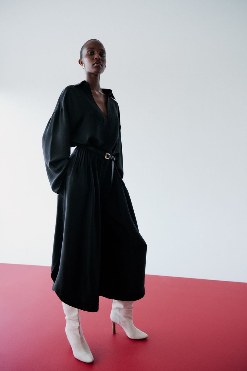 An adult female model in a black dress and white boots poses in a studio with a white and red backdrop for a fashion editorial.